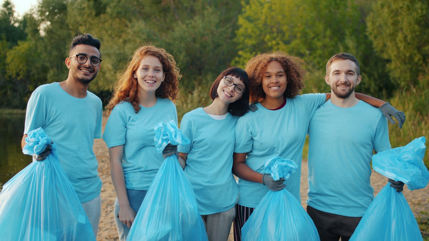Diverse volunteers smile holding blue trash bags on a path by the water.