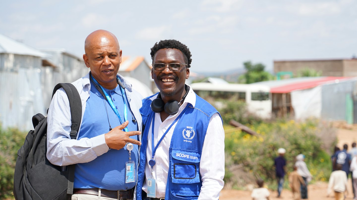 Two men wearing vests and backpacks stand near simple structures.